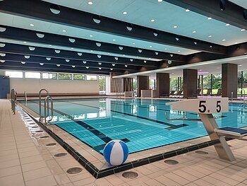 Innenaufnahme des leeren Jurabads mit Becken mit strahlend blauem Wasser. Ein Wasserball liegt dekorativ im Vordergrund.