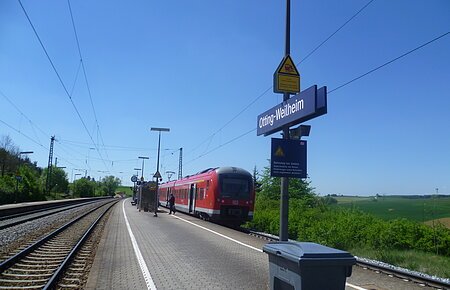 Der Bahnhof Otting-Weilheim mit rotem Zug auf dem Gleis, blauer Himmel, grüne Landschaft im Hintergrund.