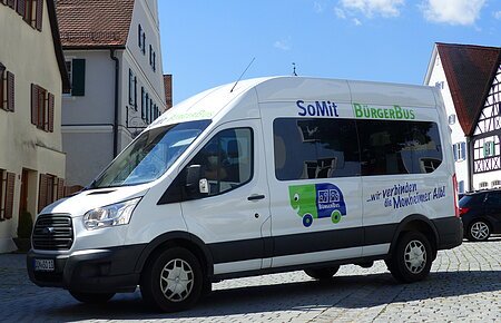 Weißer Kleinbus steht auf einem gepflasterten Platz, in der Monheimer Innenstadt . Auf dem Bus ist die Aufschrift "SoMit BürgerBus" und "wir verbinden die Monheimer Alb".