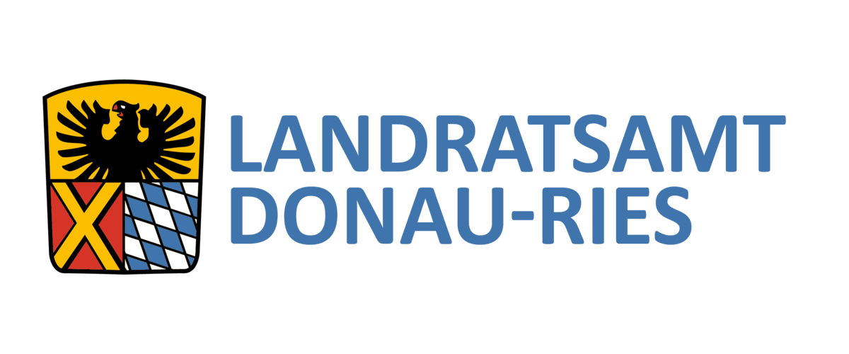 Wappen des Landratsamts DONAU-RIES: auf dem Wappen oben ein schwarzer Adler auf gelbem Hintergrund. darunter links ein gelbes Kreuz auf rotem Hintergrund und rechts davon bayerisches Rautenmuster in weiß-blau. Rechts neben dem Wappen der Text: Landratsamt Donau-Ries.