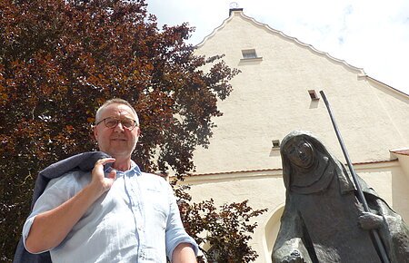 Mann mit Jacke über der Schulter steht neben einer Statue, der Walburga-Statue in Monheim vor einem gelbgestrichenem Gebäude mit Giebeldach. Im Hintergrund ein großer Baum und sonniger Himmel.