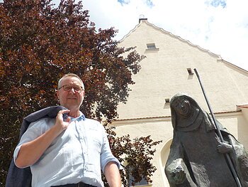 Mann mit Jacke über der Schulter steht neben einer Statue, der Walburga-Statue in Monheim vor einem gelbgestrichenem Gebäude mit Giebeldach. Im Hintergrund ein großer Baum und sonniger Himmel.