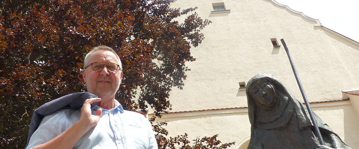 Mann mit Jacke über der Schulter steht neben einer Statue, der Walburga-Statue in Monheim vor einem gelbgestrichenem Gebäude mit Giebeldach. Im Hintergrund ein großer Baum und sonniger Himmel.