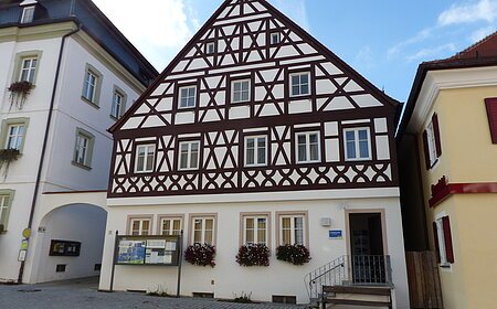 Tourist-Information der Stadt Monheim/Monheimer Alb: ein Fachwerkhaus mit drei Stockwerken, weißen Wänden und braunen Balken. Davor steht eine Sitzbank und ein Schaukasten auf einem gepflasterten Platz. Links davon ein weißes dreistöckiges Gebäude. Rechts davon ein gelbes zweistöckiges Gebäude mit roten Fensterläden.