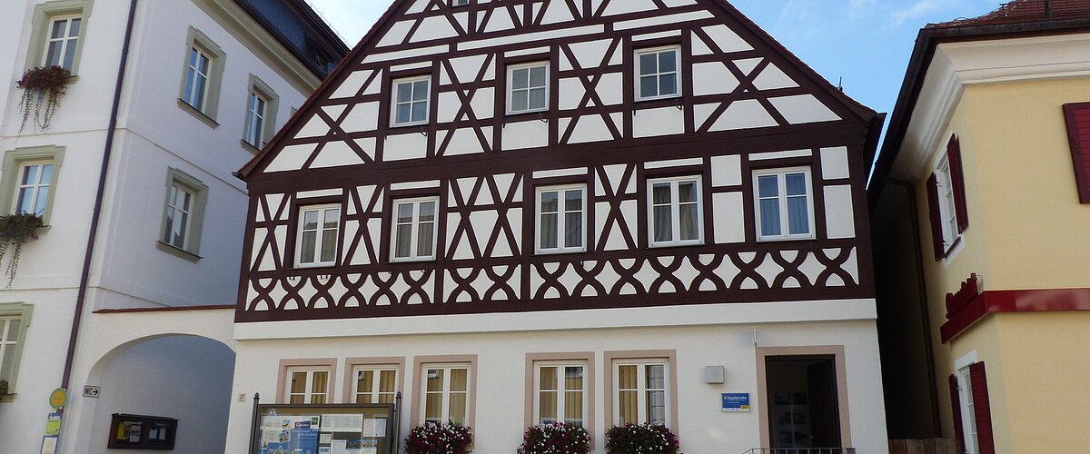 Tourist-Information der Stadt Monheim/Monheimer Alb: ein Fachwerkhaus mit drei Stockwerken, weißen Wänden und braunen Balken. Davor steht eine Sitzbank und ein Schaukasten auf einem gepflasterten Platz. Links davon ein weißes dreistöckiges Gebäude. Rechts davon ein gelbes zweistöckiges Gebäude mit roten Fensterläden.