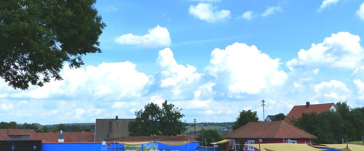 Außenbereich des Monheimer Freibads mit Schwimmbecken, Sonnensegeln, Bäumen und Gebäuden im Hintergrund unter blauem Himmel. Um das Becken grüner Rasen.