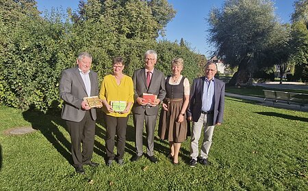 Fünf Personen stehen auf einer Wiese vor Bäumen, drei halten bunte Geschenke in den Händen, im Hintergrund blauer Himmel. Renteneintritt der drei langjährigen Mitarbeiter der VG Monheim.