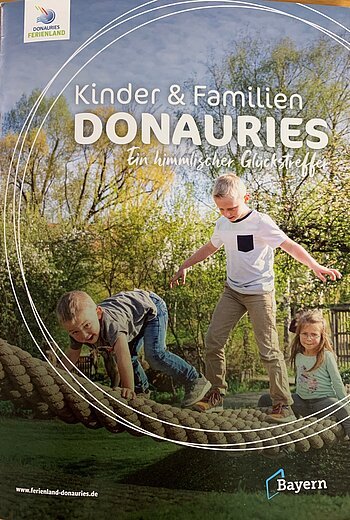 Titelseite der Broschüre "Kinder & Familien Donauries" - Ein himmlischer Glückstreffer vom Ferienland Donau-Ries. Auf dem Titelbild drei Kinder spielend auf einem Kletterseil im Freien, umgeben von Bäumen und Wiese.