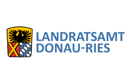 Wappen des Landratsamts DONAU-RIES: auf dem Wappen oben ein schwarzer Adler auf gelbem Hintergrund. darunter links ein gelbes Kreuz auf rotem Hintergrund und rechts davon bayerisches Rautenmuster in weiß-blau. Rechts neben dem Wappen der Text: Landratsamt Donau-Ries.