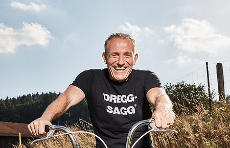 Michl Müller "Limbo of Life" Mann mit schwarzem T-Shirt mit der weißen Aufschrift "DREGG-SAGG" und Jeans sitzt lachend auf orangefarbenem Fahrrad auf Feldweg bei Sonnenschein