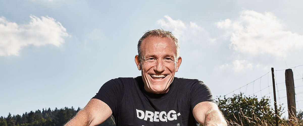 Mann mit schwarzem T-Shirt mit der weißen Aufschrift "DREGG-SAGG" und Jeans sitzt lachend auf orangefarbenem Fahrrad auf Feldweg bei Sonnenschein