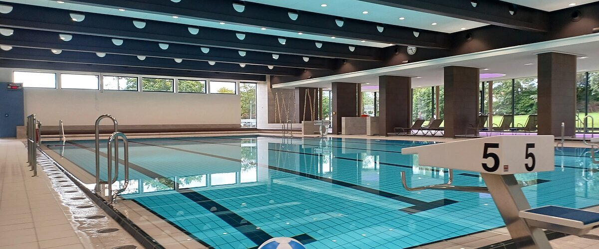 Innenaufnahme des leeren Schwimmbads - Jurabad Monheim. Zu sehen ist ein Schwimmbecken gefüllt mit strahlendem Wasser, ein Sprungbrett und ein weiß-blauer Wasserball liegt dekorativ im Vordergrund. Im Hintergrund des Hallenbads sind große Fenster zu sehen.