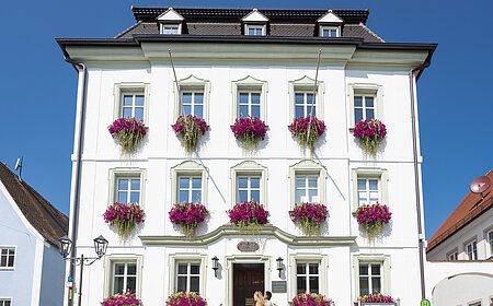 Das Rathaus in Monheim: Ein weißes dreistöckiges Gebäude mit mehreren Fenstern, an denen Blumenkästen mit pinken Blumen hängen. Zwei Personen gehen davor vorbei. Links und rechts davon jeweils ein weiteres Haus.