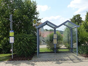 Bushaltestelle mit Glasüberdachung, umgeben von Bäumen und Sträuchern, mit Fahrplan und Haltestellenschild.