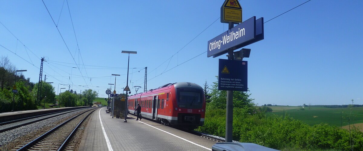 Der Bahnhof Otting-Weilheim mit rotem Zug auf dem Gleis, blauer Himmel, grüne Landschaft im Hintergrund.