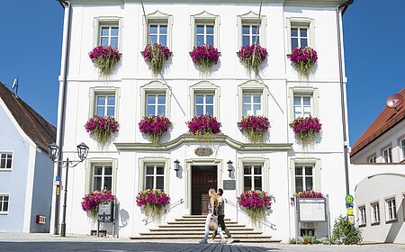 Das Rathaus in Monheim: Ein weißes dreistöckiges Gebäude mit mehreren Fenstern, an denen Blumenkästen mit pinken Blumen hängen. Zwei Personen gehen davor vorbei. Links und rechts davon jeweils ein weiteres Haus.